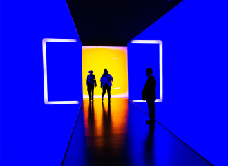 Two figures walk from a brightly lit room into a neon blue corridor, where a man waits